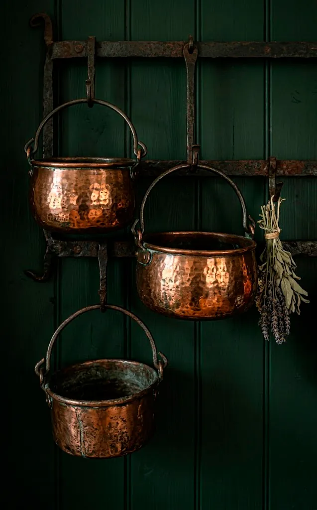 Antique hand-hammered copper jam pots with brass handles hanging on a dark green kitchen wall.