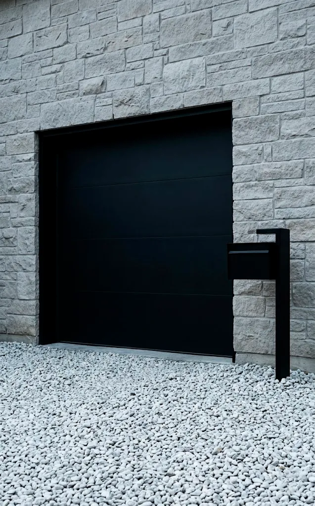 Matte black garage door against a light gray limestone wall with gravel landscaping.